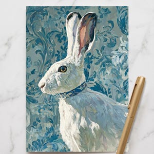 Peut inclure: Une impression rectangulaire représentant un lièvre blanc aux longues oreilles portant un collier à motifs bleus, sur un fond damassé bleu et blanc. Un stylo en métal doré est placé en diagonale dans le coin inférieur droit de l'impression.