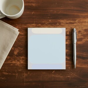May include: A square notepad with a light blue center and cream-colored top and bottom borders. A silver pen rests beside the notepad on a dark wooden surface. A white mug and beige napkin are also visible.