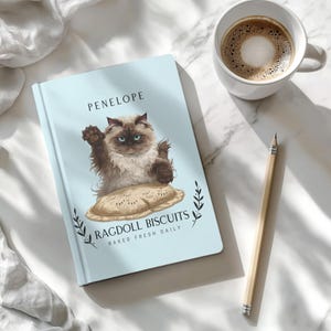 May include: A light blue notebook with the name "PENELOPE" at the top. The cover features a Ragdoll cat illustration sitting on a biscuit, with the text "RAGDOLL BISCUITS BAKED FRESH DAILY". A cup of coffee and a pencil are next to the notebook.