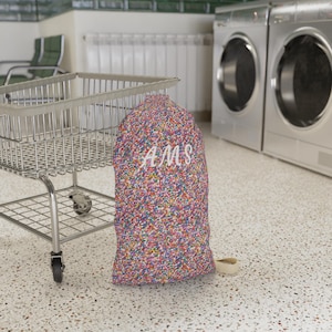 May include: A colorful laundry bag with a speckled pattern in shades of pink, blue, and yellow. The bag has the letters "AMS" in white script. A metal laundry cart is to the left, and a washing machine is in the background.