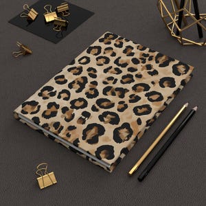 Puede incluir: Un diario de tapa dura cerrado con una cubierta con estampado de leopardo beige. Artículos de papelería dorados y negros, incluidos lápices y clips, rodean el diario. El diario parece nuevo.