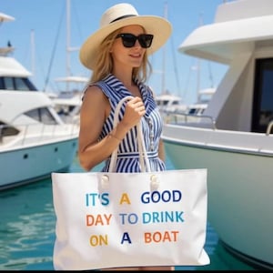 May include: A white tote bag with the text "IT'S A GOOD DAY TO DRINK ON A BOAT" in blue, orange, and yellow. The bag has white rope handles and is held in front of a woman wearing a striped dress and sunglasses. Yachts are in the background.