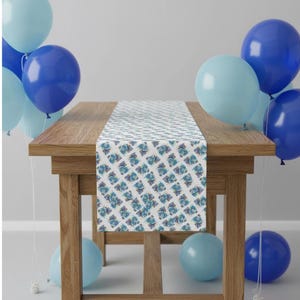 May include: A wooden table with a patterned table runner and blue balloons. The table runner has a white background with a repeating blue and teal floral design. The balloons are various shades of blue.