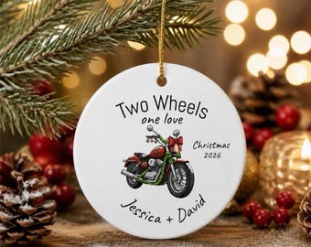 Puede incluir: Un adorno redondo de cerámica blanca con una ilustración de una motocicleta roja con una guirnalda verde. El texto dice Two Wheels one love, Christmas 2026 y Jessica + David. Tiene un cordón dorado en la parte superior para colgarlo.