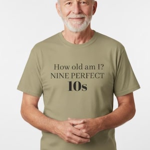 May include: A light olive green t-shirt with the text "How old am I? NINE PERFECT 10s" in black. The shirt is worn by a person with a white beard and gray hair.