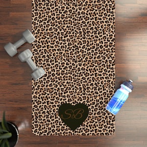 May include: A leopard print yoga mat with a dark heart-shaped design containing the letters "SB". Two gray dumbbells and a blue water bottle are also visible. The mat is designed for fitness and exercise.