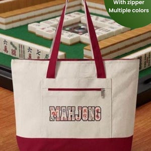 May include: Cream-colored tote bag with a red base and handles. The bag features a front pocket with a zipper and the word "MAHJONG" embroidered on the front. The bag measures 18" x 14" and has a zipper. Multiple colors are available.