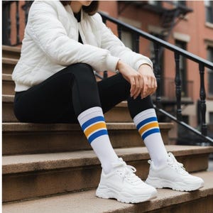 Puede incluir: Zapatillas deportivas blancas con suela gruesa combinadas con calcetines blancos con rayas azules y doradas. Los calcetines se llevan con leggings negros. También se ve una chaqueta acolchada blanca.