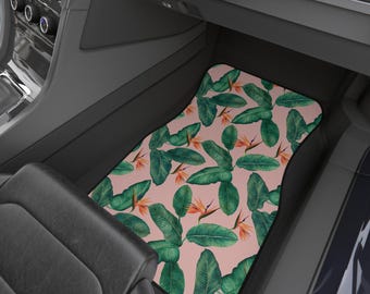 Op de afbeelding: Vloermat voor de auto met een tropisch bladerenpatroon. De mat heeft grote groene bladeren en oranje bloemen op een lichtroze achtergrond. De mat is in een auto geplaatst.