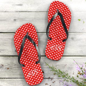 May include: A pair of red flip-flops with white polka dots and black straps. The flip-flops have a white monogram design. The sandals are on a light wood surface.
