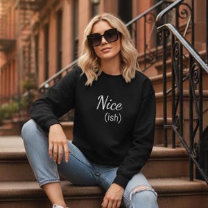May include: Black sweatshirt with the text "Nice (ish)" in white script. The person is wearing sunglasses and light blue jeans with ripped knees. The person is sitting on brown steps with a black metal railing.