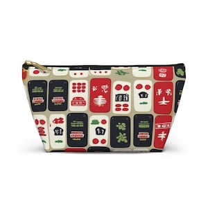 Könnte beinhalten: Eine beige Reißverschlusstasche mit einem sich wiederholenden Muster aus bunten Mahjong-Steinen. Die Steine sind in Rot-, Grün-, Schwarz- und Weißtönen gehalten und weisen verschiedene Symbole und Zeichen auf. Die Tasche hat einen goldfarbenen Reißverschluss.
