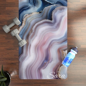 May include: A yoga mat featuring a blue, pink, and white agate pattern, with gold detailing. Two grey dumbbells and a blue water bottle are placed near the mat. The name "Sara" is printed on the mat.
