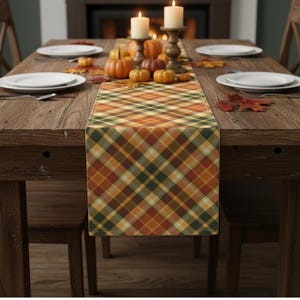 Puede incluir: Un camino de mesa rectangular con un patrón de cuadros en diagonal en tonos naranja, amarillo, marrón y verde oscuro. El camino de tela está centrado sobre una mesa de comedor de madera, decorada con pequeñas calabazas, velas y hojas.