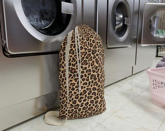 Op de afbeelding: Een waszak met luipaardprint met een trekkoordsluiting en een beige band. De tas staat rechtop voor een rij roestvrijstalen wasmachines. De tas is gevuld met wasgoed.