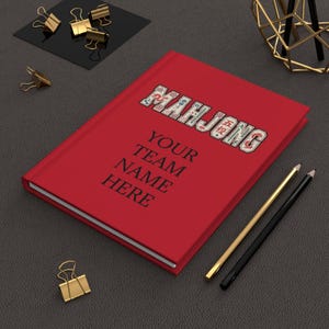 May include: A red journal with the word "MAHJONG" in a decorative font on the cover. Below the title, the text reads "YOUR TEAM NAME HERE". Gold and black pencils and gold binder clips are also visible.