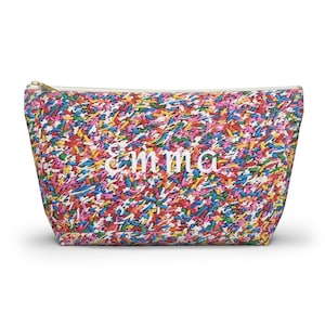 May include: A vibrant cosmetic bag featuring a sprinkle design in shades of red, yellow, blue, and pink. The name "Emma" is embroidered in white on the front. It has a white zip and a gold-coloured zip pull.