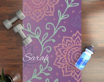 May include: A purple yoga mat with a floral mandala design in shades of pink and orange, and green vines. The name "Sarah" is written in cursive. Two gray dumbbells and a blue water bottle are also present.