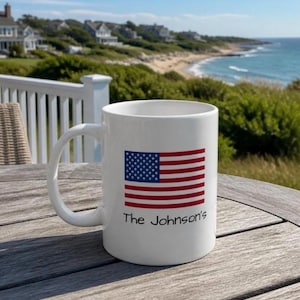 May include: A white ceramic mug featuring a printed graphic of the United States flag with the text The Johnson's positioned directly underneath the flag. The mug has a curved handle and a smooth finish, displayed on a wooden surface.