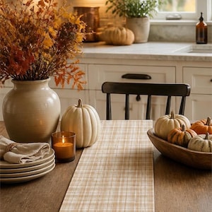 May include: A rectangular table runner featuring a light tan and white plaid pattern centered on a dark wooden dining table. The runner is accompanied by a ceramic vase filled with autumn foliage, several small pumpkins, and a stack of plates.