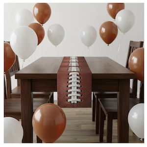 May include: A brown table runner with a football design, featuring white laces, is centered on a dark wooden table. White and brown balloons are arranged around the table, creating a festive atmosphere. Wooden chairs are placed around the table.