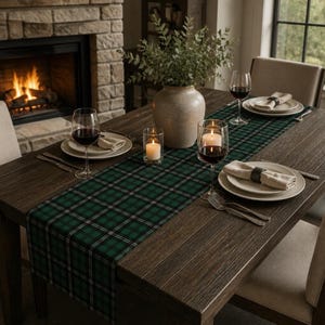 May include: A dark wooden dining table set with a green and black plaid table runner, white ceramic plates, beige cloth napkins with dark napkin rings, clear wine glasses filled with red wine, two lit pillar candles, and a vase with greenery.