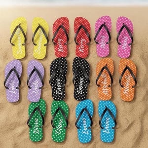 May include: A collection of personalized flip-flops in various colors, including yellow, red, pink, black, orange, green, purple, and blue. Each flip-flop features a polka dot pattern and a name printed on the sole. The flip-flops are arranged on a sandy surface.
