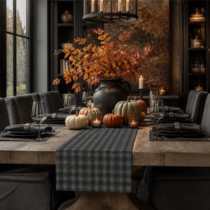 May include: A wooden dining table featuring a grey plaid table runner, a large black vase filled with orange autumn foliage, and an assortment of white, orange, and green pumpkins. The table is set with black plates, napkins, and wine glasses.