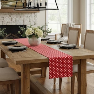 Puede incluir: Una mesa de comedor de madera con un camino de mesa de lunares rojos y blancos. La mesa está puesta con platos negros y cubiertos. Un jarrón de flores blancas está en el centro. La habitación tiene una chimenea y una lámpara de araña.