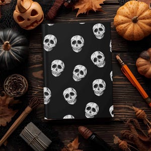 May include: A black journal with a repeating pattern of white skulls. The journal is surrounded by Halloween-themed decorations, including pumpkins, leaves, and a pencil, all arranged on a dark wooden surface.