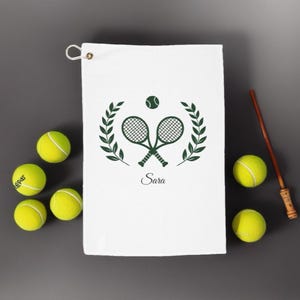 May include: White tennis towel with a green tennis racket and laurel wreath design, and the name "Sara" printed below. Several yellow tennis balls surround the towel. A wooden stick with a decorative handle is also present.