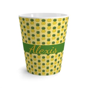 Puede incluir: Una taza de café con leche de cerámica blanca con un patrón repetido de piñas amarillas sobre un fondo amarillo claro. Una banda verde con el nombre "Alexis" en cursiva envuelve el centro de la taza.