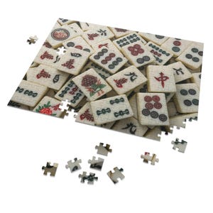 May include: A partially assembled Mahjong puzzle with ivory-colored tiles. The tiles feature various symbols, including Chinese characters, circles, and floral designs. Several puzzle pieces are scattered around the main section.