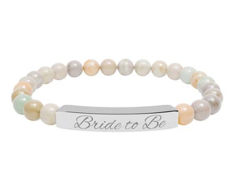 May include: A beaded bracelet with a mix of pastel-colored beads. The bracelet features a silver-colored bar with the engraved text "Bride to Be". The beads are in shades of cream, peach, gray, and green.