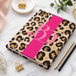 May include: A hardback journal featuring a tan and black leopard print pattern with a wide, vertical hot pink stripe running down the centre. The letters BD are printed in a large, stylised light pink font over the centre of the stripe.