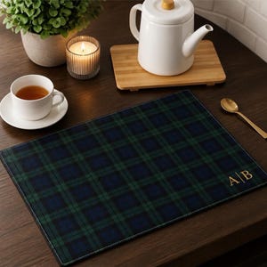 May include: A rectangular placemat featuring a dark green and navy blue plaid pattern with a gold embroidered monogram of the letters A and B separated by a vertical line in the bottom right corner, finished with a thin black stitched border.