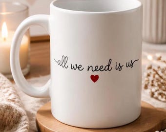May include: A white ceramic mug featuring the black cursive text "all we need is us" centered on the front, positioned above a small, solid red heart icon. The mug has a smooth, cylindrical body with a curved handle on the left side.