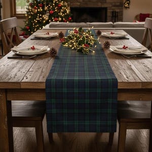 May include: A wooden dining table set for a holiday meal. A blue and green plaid table runner runs down the center, adorned with holly and lights. Plates, silverware, and pinecones are arranged for each place setting. A Christmas tree is visible in the background.