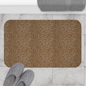 May include: A rectangular bath mat with a leopard print pattern in shades of brown and tan. The mat is placed on a tiled floor, with a pair of gray slippers and a rolled-up gray towel visible.