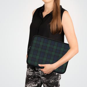 Op de afbeelding: Een rechthoekige laptoptas met een zwart, groen en blauw ruitpatroon. De hoes heeft een zwarte rits en rand. Het product wordt vastgehouden door een persoon die een zwarte mouwloze top en camouflagebroek draagt.