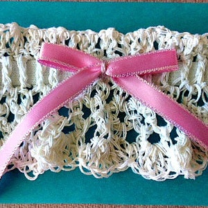 May include: A cream-colored crocheted garter featuring an intricate openwork pattern and a central elastic band. A smooth, satin-finish pink ribbon is tied into a bow at the center of the garter, resting against a solid teal rectangular base.