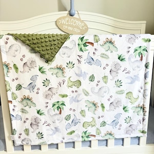 May include: A white baby blanket featuring a repeating pattern of light green and grey dinosaurs, palm trees, and leaves. The blanket has a textured olive green minky fabric backing that is folded over at the top corner to show the underside.