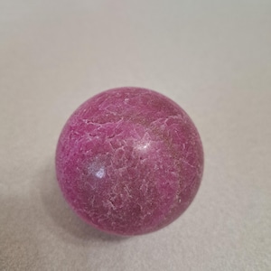 May include: A spherical, polished gemstone in a deep magenta hue. The surface has a textured, cracked appearance, with subtle variations in color. The sphere is a solid, opaque object, likely used for decorative or metaphysical purposes.