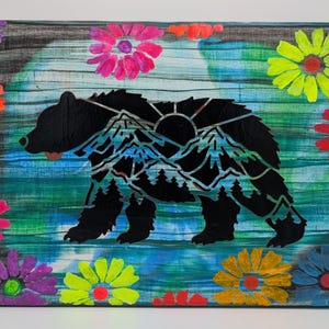 May include: A rectangular canvas painting featuring a black bear silhouette filled with a mountain range and forest landscape. The background consists of horizontal blue and green brushstrokes, framed by vibrant, multi-colored painted flowers.