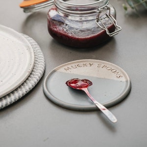 May include: A grey and white ceramic dish with the words "Mucky Spoons" printed on it. A silver spoon with a dollop of red jam is on the dish.