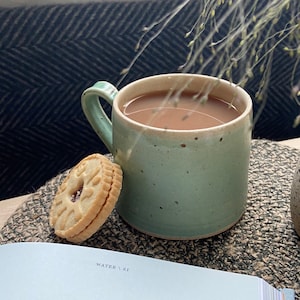 May include: A light green ceramic mug with a speckled glaze filled with brown tea. A biscuit with a red filling sits on a woven placemat next to the mug.
