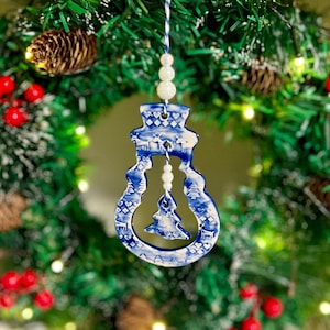 May include: A blue and white ceramic Christmas ornament in the shape of a lightbulb, with a small Christmas tree inside. The ornament is hanging from a white and blue string, and is surrounded by a green wreath with red berries and pinecones.