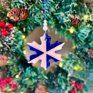 May include: A white ceramic snowflake ornament with blue paint, hanging from a string of white beads on a green Christmas tree.