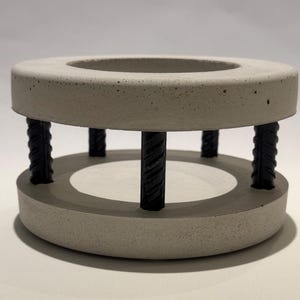 Könnte beinhalten: Grauer Beton-Kerzenhalter mit kreisförmigem Design. Der Halter hat einen oberen und unteren Ring, die durch schwarze, strukturierte Metallstäbe verbunden sind. Die Mitte des oberen Rings ist vertieft, um eine Kerze aufzunehmen. Modernes, industrielles Design.