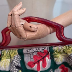 Peut inclure: Un sac &agrave; main vintage avec une poign&eacute;e rouge et un corps en tissu &agrave; motifs. Le tissu pr&eacute;sente des feuilles rouges, des ananas et d'autres motifs tropicaux sur un fond vert. La poign&eacute;e est en plastique rouge transparent incurv&eacute;.